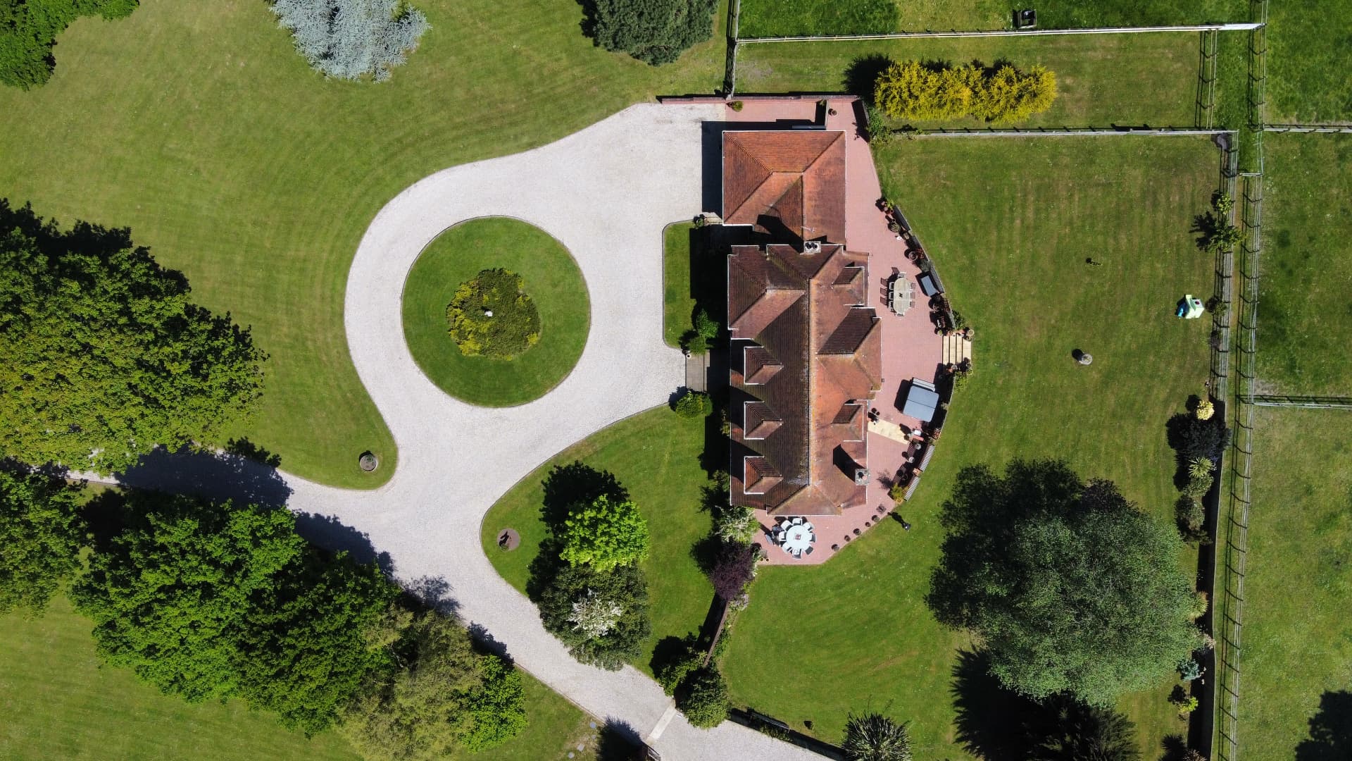 An aerial view directly overhead of a country house with a circular gravel drive and lawned grounds bordered by mature trees